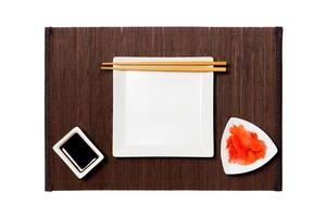 Empty white square plate with chopsticks for sushi, ginger and soy sauce on dark bamboo mat background. Top view with copy space for you design photo