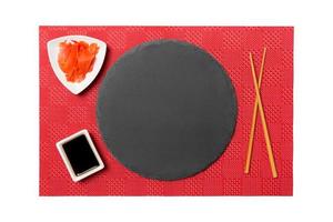 Emptyround black slate plate with chopsticks for sushi and soy sauce, ginger on red mat sushi background. Top view with copy space for you design photo