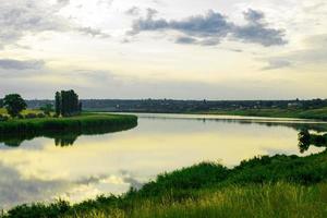 Summer evening dawn sky clouds reflected on a lake of water on the coast with green grass and forest trees photo