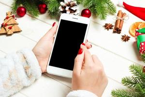 Top view of a woman holding a phone in her hand on wooden New Year background made of fir tree and festive decorations. Christmas holiday concept. Mockup photo