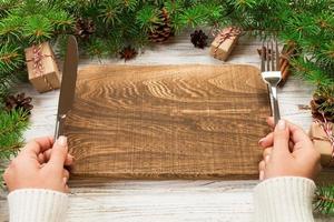 Perspective view girl holds fork and knife in hand and is ready to eat. Empty wood rectangular plate on wooden christmas background. holiday dinner dish concept with new year decor photo