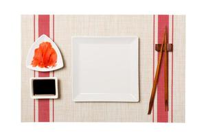 Empty white square plate with chopsticks for sushi and soy sauce, ginger on sushi mat background. Top view with copy space for you design photo