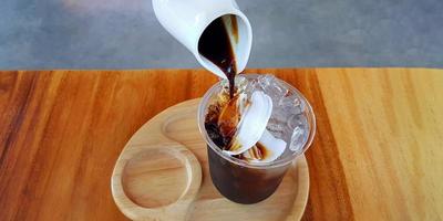 Pouring black or dark coffee in plastic glass of ice with fresh coconut on brown wooden table at caf. Cold drinking and refreshment. Iced coconut coffee menu. photo