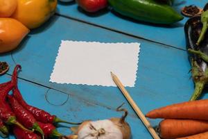 Vegetables are laid out around a sheet of paper and a pencil. Empty space for text. Vegetables, empty blank for recipe on a blue background. photo