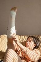 girl with an expression of pain on her face, lies on the couch with her leg raised up in a cast. The girl corrects the plaster on her leg or doing exercises photo