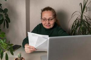 A middle-age businesswoman works at a desk, makes notes in a laptop, and checks the data on her phone. Business, online business, work in the office photo