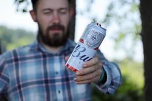 KHARKOV, UKRAINE - MAY 5, 2022 Young man raise Budweiser Bud beer can and shows BUD logo on blurred river and trees background. Budweiser is one of the most popular beer brands in the USA photo