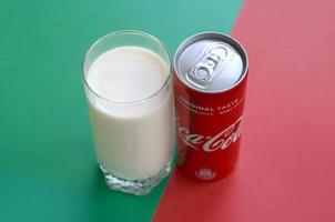 KHARKOV. UKRAINE - MAY 2, 2022 Coca cola red tin can and cup of fresh milk on red and green background surface photo