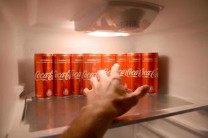 KHARKOV. UKRAINE - MAY 2, 2022 Male hand want to grab Coca-cola red drink can inside domestic cooler fridge full of coke cans photo