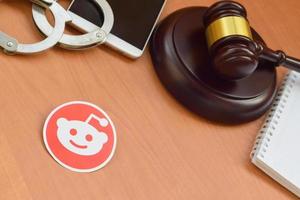 KHARKOV, UKRAINE - MAY 12, 2022 Reddit paper logo lies with wooden judge gavel, smartphone and handcuffs. Entertainment lawsuit concept photo