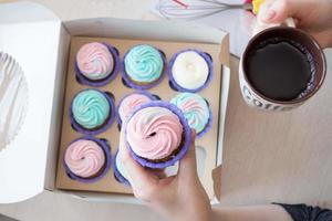 cupcake with pink and blue cream in the hands of a girl on the background of a box of cupcakes and cup of coffee photo