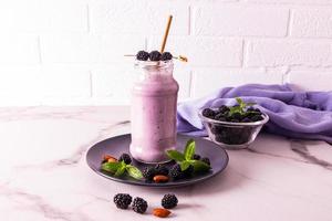 A glass bottle of blackberry smoothie or cocktail stands on a dark ceramic plate on a marble table against a brick wall. healthy eating. diet. photo