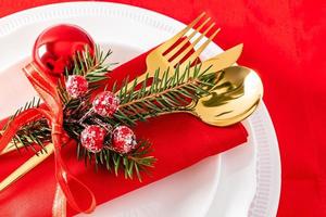 the near view of part of the plates serving the New Year's table. gold cutlery with a spruce branch and red balls on a red background.