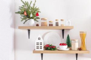 wooden open kitchen shelves on a white textured wall with cans for loose products, a bouquet of spruce branches, balls and a cardboard white house. photo