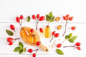 The top view of several glass bottles with a cork with natural rosehip seed oil lie on a wooden board and a white wooden table among the ripe fruits. photo