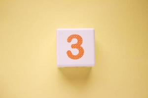 Close-up photo of a white plastic cube with a orange number 3 on a yellow background. Object in the center of the photo