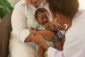 Newborn baby one-month-old mixed race African-Thai, crying sick visiting the doctor for checkup health with stethoscope at clinic pediatric. concept consultation, check-up, nurse, healing infant. photo