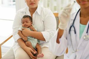 Mother holding newborn baby boy one-month-old mixed-race African-Thai, visiting the doctor for checkup health with stethoscope at the clinic pediatric. concept consultation, nurse, healing infant. photo