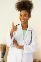 retrato de una joven doctora afroamericana de pie, feliz, sonriente, confiada, orgullosa y positiva, y pulgares hacia arriba mirando la cámara. consulta clínica concepto de salud y medicina. foto