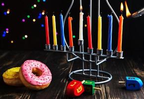 Jewish holiday Hanukkah background. A traditional dish is sweet donuts. Hanukkah table setting candlestick with candles and spinning tops on black background. Lighting Chanukah candles. Copy space photo