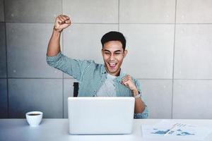 Small Business and Successful Concept. Young Asian Businessman Glad to recieve a Good News or High Profits from Computer Laptop, Own Business Achieves Goals. Raising Arms to Celebrating Success photo