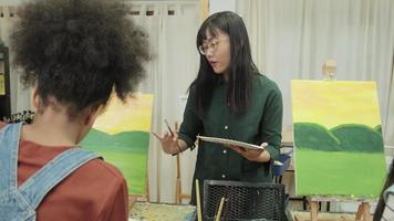 A female Asian teacher teaches and demonstrates to the children on acrylic color picture painting on canvas in art classroom, creatively learning with skill at the elementary school studio education. video