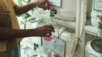 Close-up shot of customer's hands filling natural products in recycled bottle from reusable containers in zero-waste and organic refill store. Thai text labels mean foam soap and dish washing liquid. video