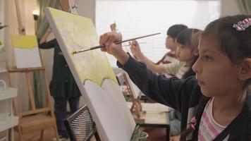 uma garotinha se concentra na pintura acrílica colorida sobre tela com crianças multirraciais em uma sala de aula de arte, aprendizado criativo com talentos e habilidades na educação de estúdio do ensino fundamental. video