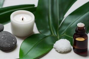 artículos de tratamiento de belleza para procedimientos de spa en mesa de madera blanca con planta verde. piedras de masaje, aceites esenciales y sal marina con velas encendidas. foto