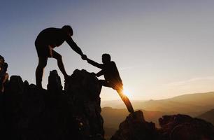 silueta de dos personas escalando el acantilado de la montaña y una de ellas dando la mano. gente ayudando y, concepto de trabajo en equipo. foto