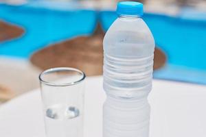 Glass and bottle with a water on white table agains swimming pool background photo