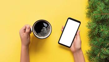 Female hands holding black mobile phone with blank white screen and mug of coffee. Mockup image with copy space. Top view banner on yellow background, flat lay photo