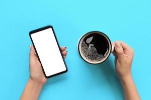 Female hands holding black mobile phone with blank white screen and mug of coffee. Mockup image with copy space. Top view on blue background, flat lay photo