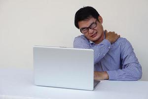 un hombre asiático se siente herido en el cuello y el hombro durante mucho tiempo trabajando en una computadora portátil. concepto, síndrome de oficina. problema de salud. auto masaje para el alivio. trabajo duro foto