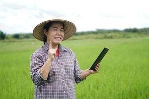 Asian woman  is at green paddy field, wears hat and plaid shirt, holds smart tablet. Point finger up.Smiles. Concept, agriculture occupation, using wireless technology device to research. Smart farmer photo