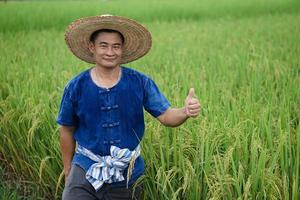 Asian man farmer is at paddy field, wears hat, blue shirt, thumbs up, feels confident.  Concept  Agriculture occupation. Working with nature. Organic farming. photo