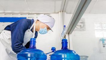 inspección de control de calidad. trabajador en ropa de trabajo y con una máscara protectora en la cara trabajando en una fábrica de agua potable revisando los galones de agua antes del envío. foto