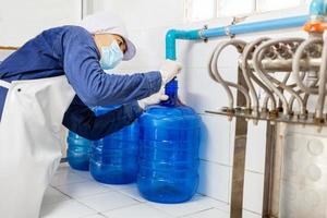 hombre trabajador en ropa de trabajo y con una máscara protectora en la cara trabajando produce agua potable en una fábrica de agua potable limpia. línea de producción de agua potable limpia foto