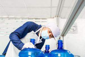 inspección de control de calidad. trabajador en ropa de trabajo y con una máscara protectora en la cara trabajando en una fábrica de agua potable revisando los galones de agua antes del envío. foto