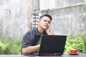 Un joven asiático atractivo que usa una laptop confundido en un espacio de trabajo conjunto con una cara infeliz foto