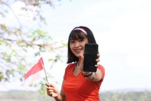 hermosa joven asiática que lleva la bandera indonesia con una cara alegre y llama a su amigo foto