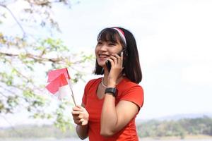 hermosa joven asiática que lleva la bandera indonesia con una cara alegre y llama a su amigo foto