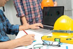 Asian engineer and partner team learn and talk with house plan project blueprint, graph and yellow helmet and engineering construction tools on table work. photo