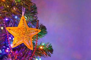 estrella amarilla decoración navideña hecha a mano con adorno en el abeto en la noche de vacaciones, colorida guirnalda de bokek, cuentas rojas, fondo púrpura oscuro borroso. tarjeta de felicitación de hadas de Navidad con espacio de copia. foto
