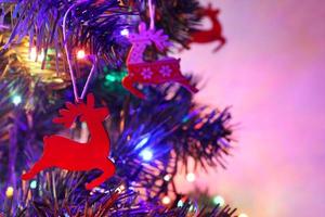 Christmas greeting card with three red santa's reindeers, one close up. Traditional wooden decorations with ornament on Christmas fir tree. Colorful bokeh garland, blurred background, purple backlight photo