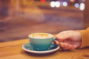 mano de hombre con una taza de café en un café sobre un fondo de luces de la ciudad detrás de la ventana. foto tonificada.