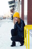 Young woman with blue eyes and blond hair in a yellow knitting hat and black leather jacket on the snowy street of city. photo