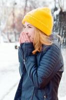 atractiva mujer joven con cabello rubio en un sombrero de punto amarillo está calentando sus manos en el parque de invierno. foto