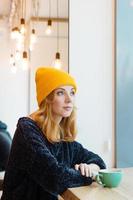 Young woman with blue eyes and blond hair in a yellow knitting hat is drinking coffee in a cafe and looking into the window. photo