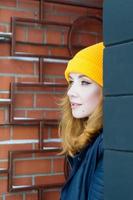 Beautiful young woman with blonde hair and blue eyes in a yellow knitting hat is looking from the corner on a background of red brick wall. Female city portrait. photo